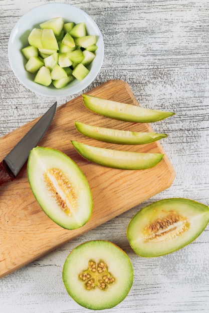 Sliced melon in a wooden cutting board with melon in bowl and knife flat lay on a white stone background