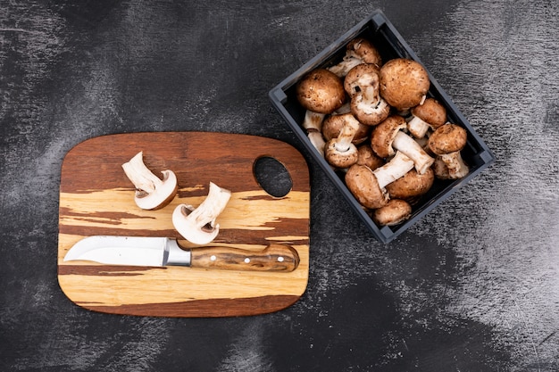 Sliced mushroom with a knife on wooden cutting board near wooden box with fresh mushrooms