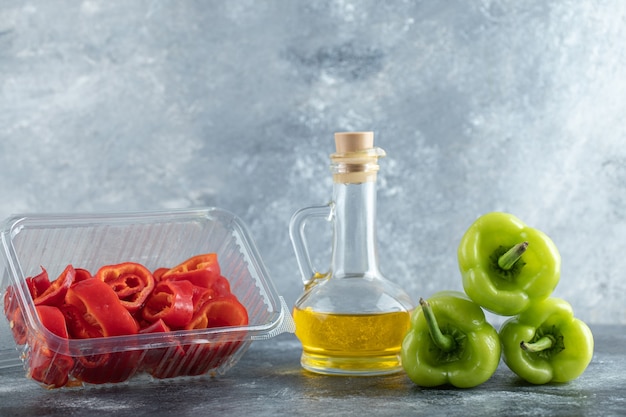 Sliced red pepper in plastic container with green pepper and bottle of oil on grey background