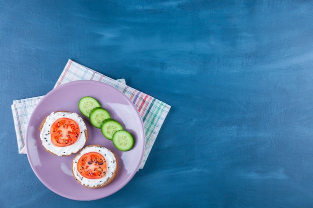 Sliced tomatoes on cheese bread next to cucumber on plate on tea towel, on blue. 