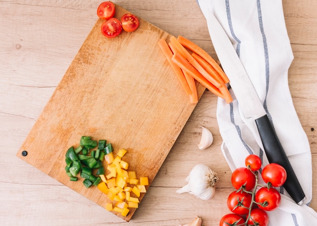 Free Photo slices of bell peppers; carrot; halved tomatoes on chopping board with garlic bulb; knife and napkin over the wooden desk