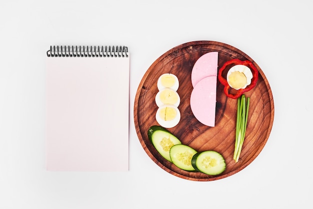 Free Photo slices of boiled egg, sausage, green onion, red pepper and cucumbers on wooden plate with notebook aside.