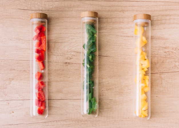 Free photo slices of red; green and yellow bell pepper slices in the transparent test tubes container on wooden desk