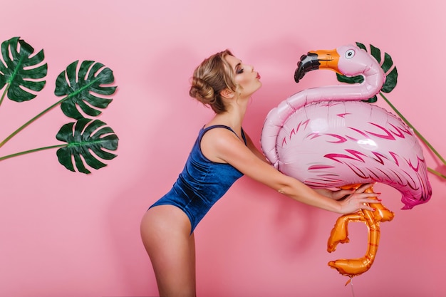 Free photo slim amazing girl in vintage bodysuit kissing big toy bird, standing in front of pink wall. portrait of cute shapely young woman holding inflatable flamingo, posing with plants on background