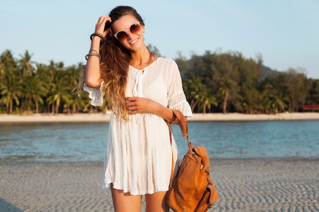 Free photo slim beautiful woman in white dress on tropical beach on sunset holding leather backpack.