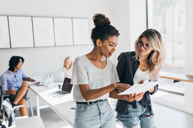 Free Photo slim black woman in jeans explaining something to european female colleauge while asian man talking with fair-haired young lady. portrait of managers of international company solving work problems.