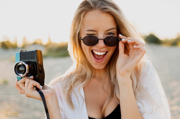 Slim blond  happy woman  holding retro camera and having fun on warm sunny beach. Summer vacation and travel concept. Natural beauty, vacation in Asia. Trendy sunglasses, white outfit.