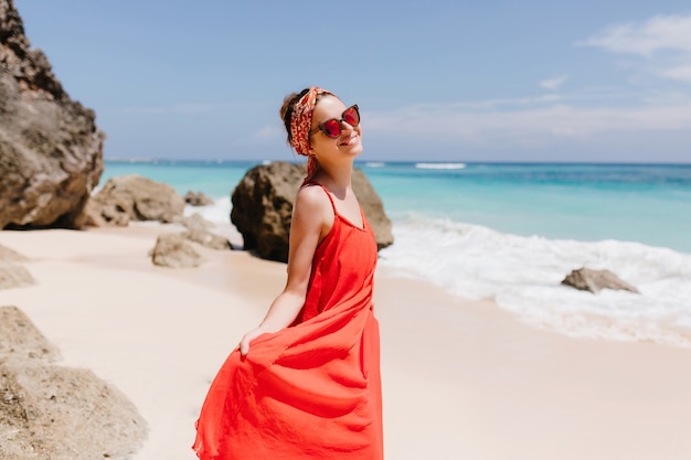 Slim carefree girl enjoying summer weekend at wild beach. Outdoor photo of adorable laughing lady in red dress standing on beach