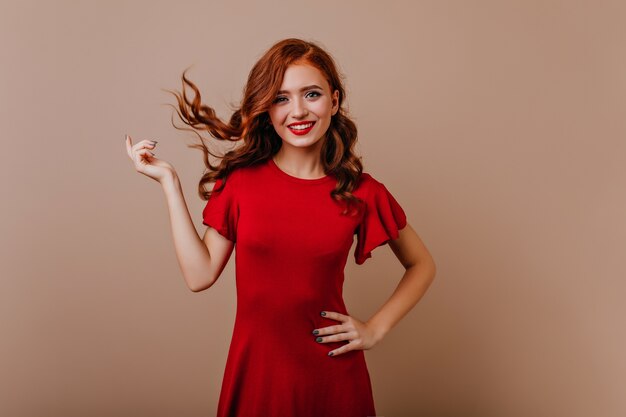 Slim caucasian woman in red dress laughing . Gorgeous ginger girl playing with her hair during photoshoot.