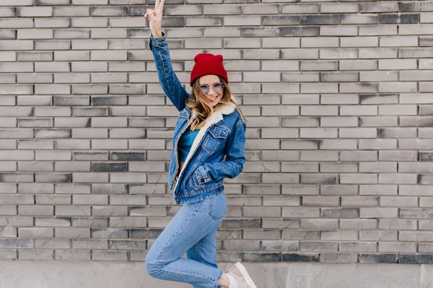 Slim girl in trendy jeans having fun on the street in cold spring day. Blissful female model in denim attire dancing on urban wall and waving hands.