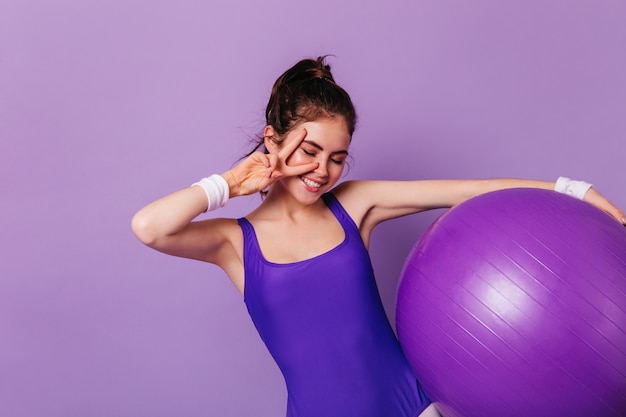 Slim gymnast woman holds fitball and shows peace sign on purple wall