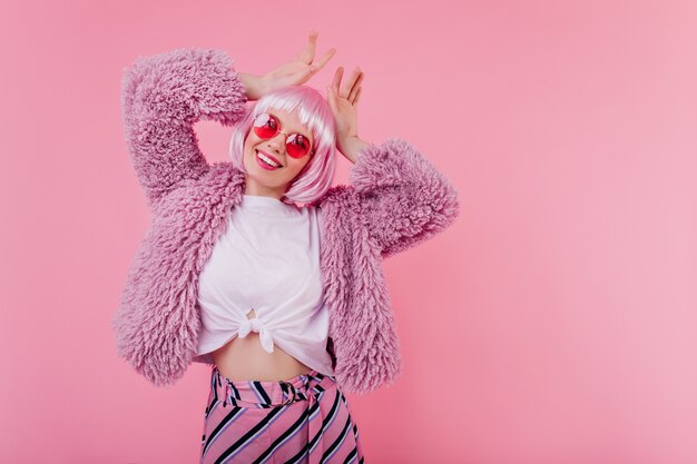 Slim lovely girl in fluffy jacket fooling around on pink wall. Interested young woman in stylish peruke expressing happiness during photoshoot