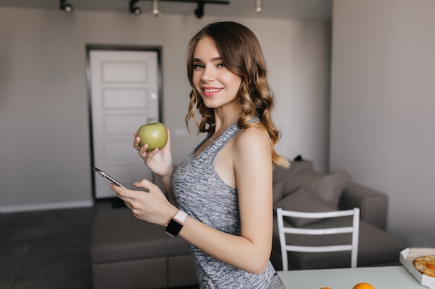 Free photo slim pale girl in smartwatch posing at home. charming dark-haired lady with apple having fun in morning.