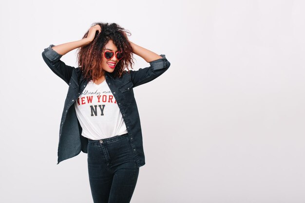 Slim smiling girl wears jeans and sunglasses dancing in studio enjoying photoshoot. Young african female model playing with her curly hair and laughing on white background.