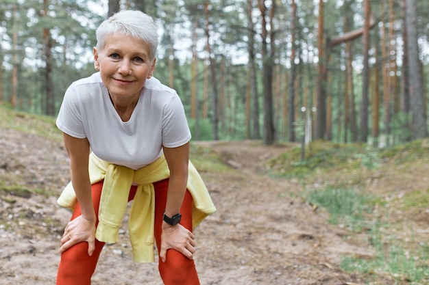 Slim sporty middle aged woman in activewear standing on pine trees background leaning forward