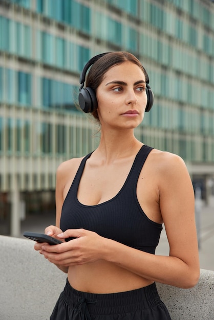 Free photo slim sporty woman looks away pensively uses mobile phone for chatting listens music via headphones dressed in active wear has regular morning training in urban setting. people and sport concept