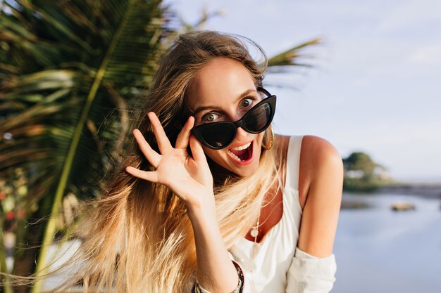 Slim woman enjoying in exotic place. Joyful tanned lady standing near palm trees and smiling.