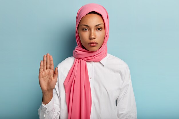 Slow down and stop. Serious dark skinned Arabic woman shows refusal gesture, raises palm towards, expresses refusal, wears pink headwear and white shirt, isolated over blue wall.