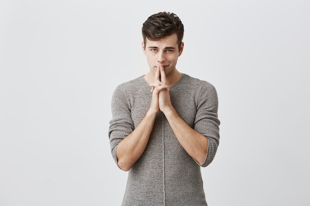 Sly man looking with cunning eyes holding his hands together trying to ask something in sly manner. Handsome young male with dark hair dressed casually expressing slyness in front of camera