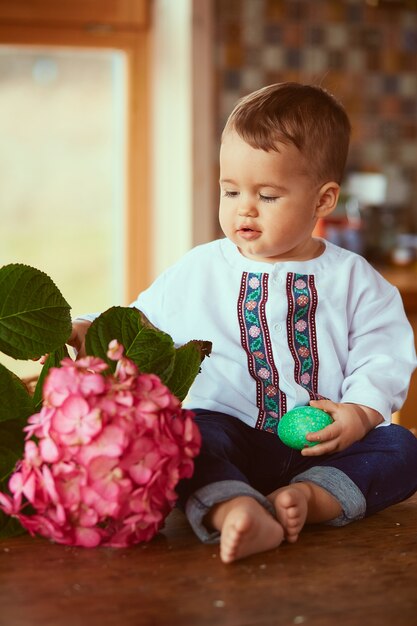 The small baby keeps a green egg and sits near flower