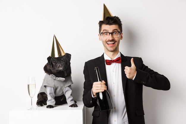 Small black dog wearing party hat and standing near happy man celebrating holiday, owner showing thumb-up and holding champagne bottle, white background