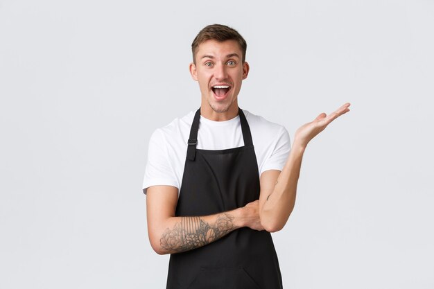 Small business owners, coffee shop and staff concept. Relieved and happy smiling handsome barista rejoicing, raise hand and looking excited, telling big news, advertising.