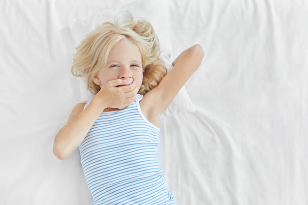 Free photo small child with blonde hair, blue eyes and freckled skin, lying in bed, covering mouth with hand and yawning. adorable little girl waking up in morning, having sleepy expression after sleep