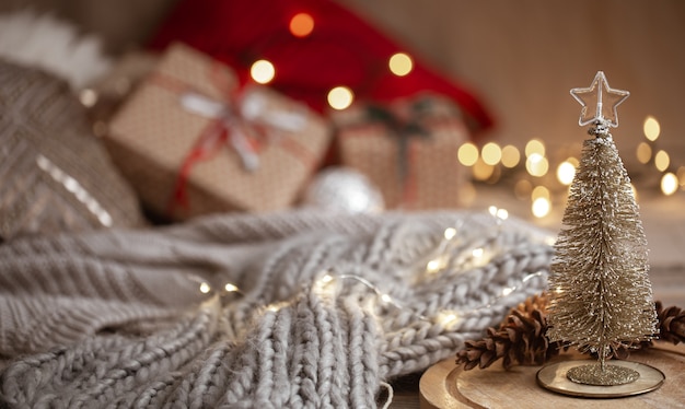 Small decorative shiny Christmas tree in the foreground on a blurred background of a knitted scarf, Christmas decorations and bokeh lights copy space.