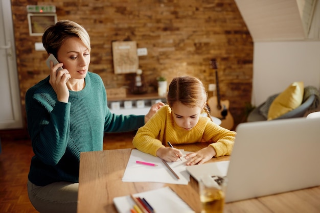 Small girl drawing on the paper while her mother is talking on the phone at home