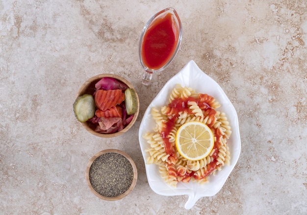 Small pasta platter, lemon slice, assorted pickles and a bowl of black pepper on marble surface.