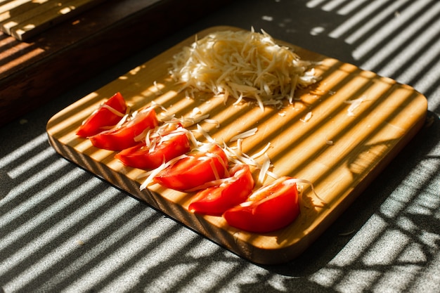 A small pile of grated fresh cheese and red tomatoes lies on a wooden board in the kitchen