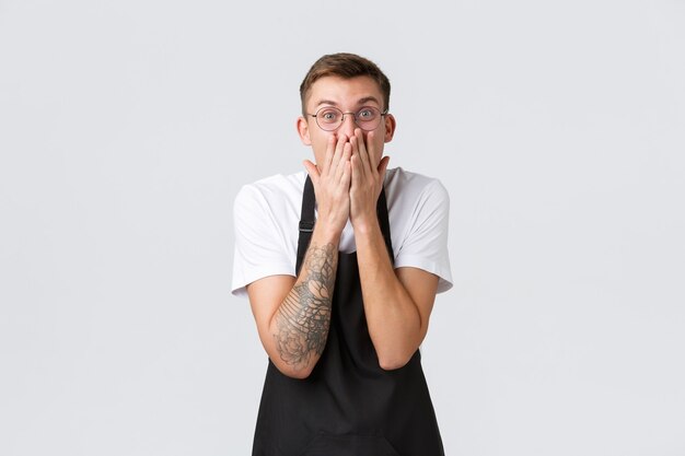 Small retail business owners, cafe and restaurant employees concept. Excited and amazed happy waiter react to amazing news, hold hands over mouth and smiling amused, standing white background