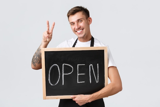 Small retail business owners, cafe and restaurant employees concept. Friendly happy barista, salesman showing peace sign and we are open sign, inviting clients enjoy coffee, white background