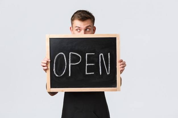 Free photo small retail business owners, cafe and restaurant employees concept. silly handsome barista, salesman looking away, holding open sign near face, standing white background