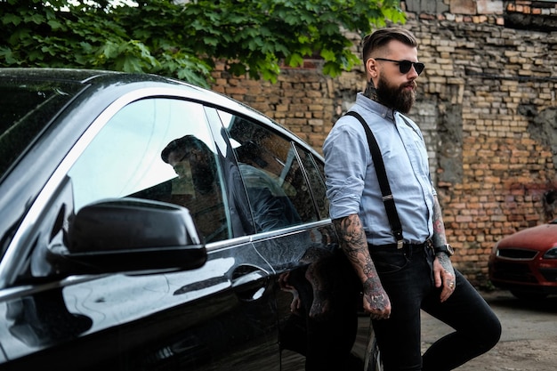 Smart bearded man is leaning on his car. He is wearing shirt, suspender and sunglasses. He has tattoes and watch on his arms. There are slum and tree at the background.