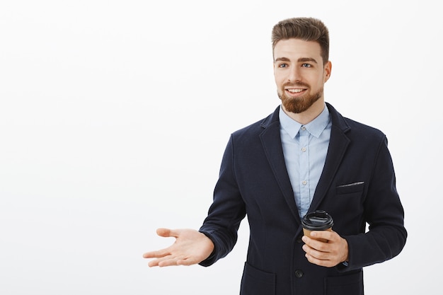 Smart and creative charismatic male entrepreneur in stylish suit holding paper cup of coffee during break talking with business partner discussing work and money gesturing with palm smiling assured