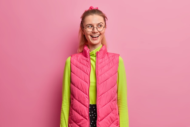 Free photo smart good looking girl being in great mood, smiles positively, looks aside, watches hilarious show, dressed in bright clothes, optical glasses