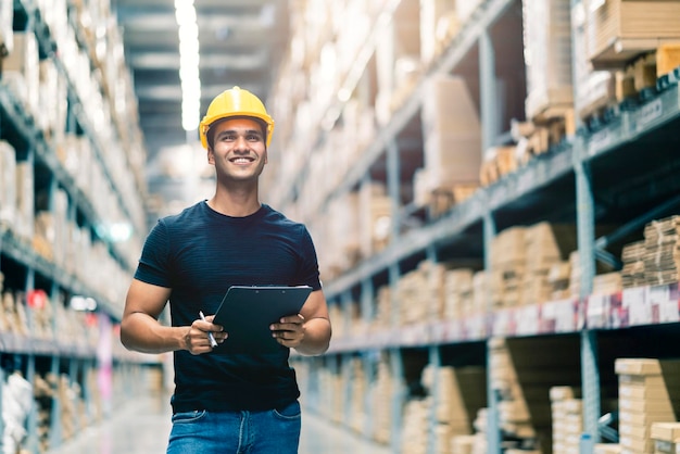 Smart Indian engineer man wearing safety helmet doing stock tick check and cardboard stock product management in factory warehouse background