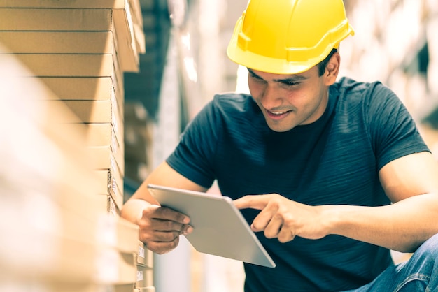 Smart Indian engineer man wearing safety helmet doing stock tick check and cardboard stock product management in factory warehouse background