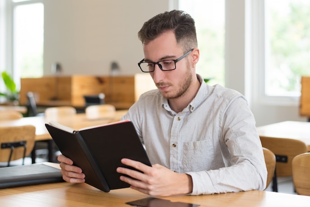 Free photo smart male student reading textbook at desk in classroom