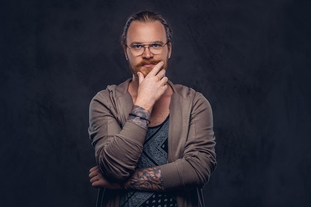 Smart pensive redhead hipster with full beard and glasses dressed in casual clothes, poses with hand on chin in a studio. Isolated on the dark background.