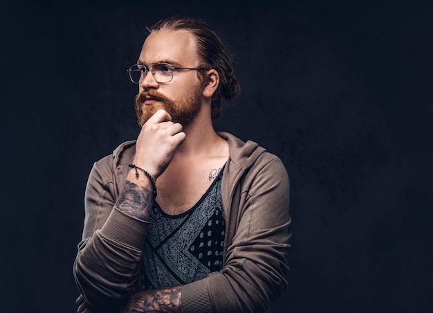 Smart pensive redhead hipster with full beard and glasses dressed in casual clothes, poses with hand on chin in a studio. Isolated on the dark background.