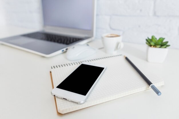 Smartphone, notebook and pencil in front of laptop on white desk