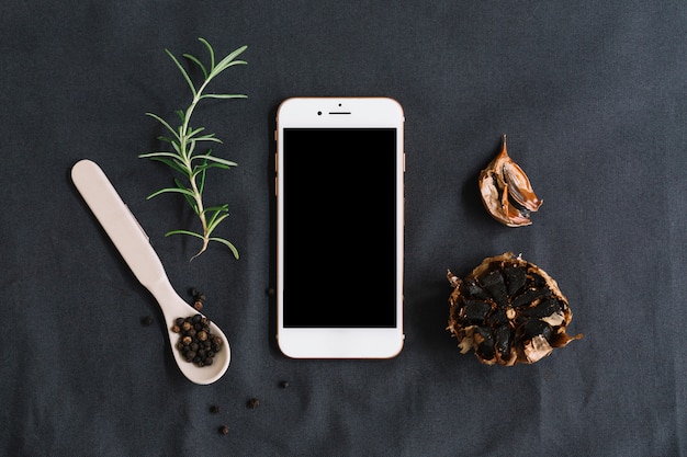 Free photo smartphone surrounded with rosemary; black pepper and garlic on dark backdrop