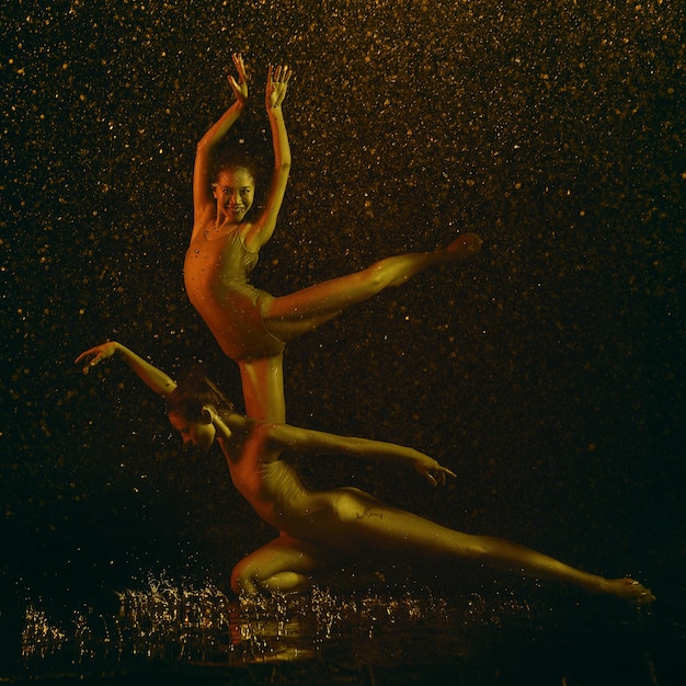 Free photo smile. two young female ballet dancers under water drops and spray. caucasian and asian models dancing together in neon lights. ballet and contemporary choreography concept. creative art photo.