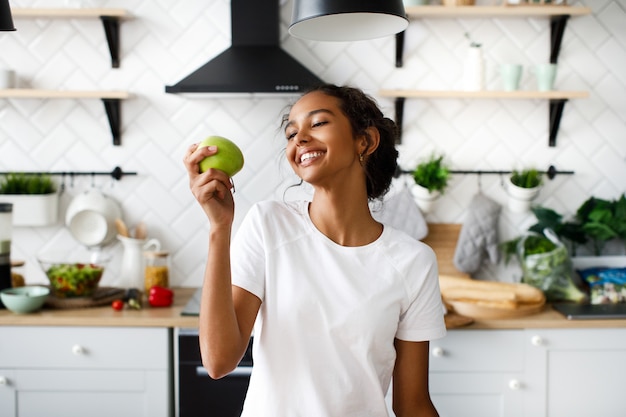 Smiled attractive mulatto woman is preparing to bite an apple and looking on apple on the white modern kitchen