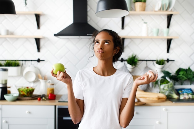 Smiled attractive mulatto woman is thinking about an apple with hilarious face and looking to the top on the white modern kitchen