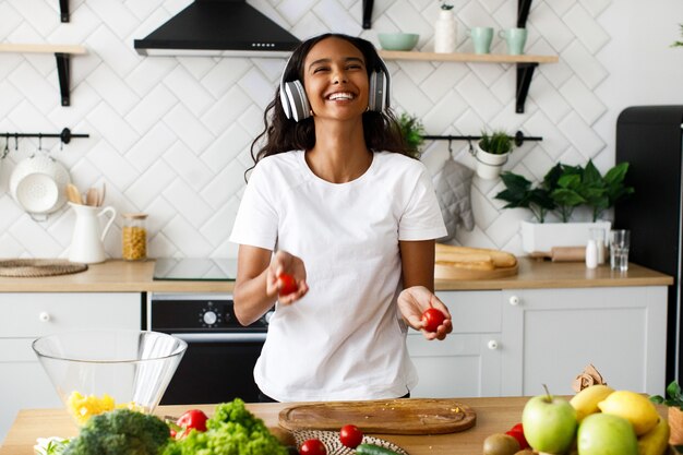 Smiled beautiful mulatto woman is holding tomatoes and listening something in big headphones near the table full of the fresh vegetables on the modern kitchen dressed in white t-shirt