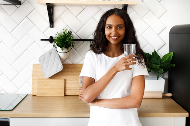 Smiled mulatto woman is holding glass with water near the kitchen desk on the modern white kitchen