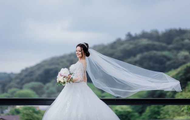 Smiled pretty caucasian bride with long veil outdoors holding wedding bouquet